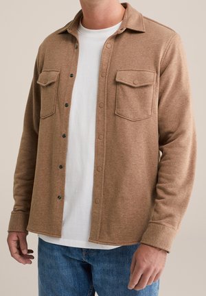 Hombre con camisa chaqueta marrón abotonada sobre una camiseta blanca y jeans azules frente a un fondo liso.