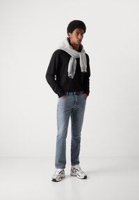Zwarte longsleeve, grijze trui over de schouders gedrapeerd, lichtblauwe jeans en witte sneakers met zwarte accenten. Effen achtergrond.