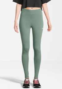 Vrouw in hoge taille saliegroene leggings en zwarte sportieve schoenen met roze veters, standing tegen een witte achtergrond.