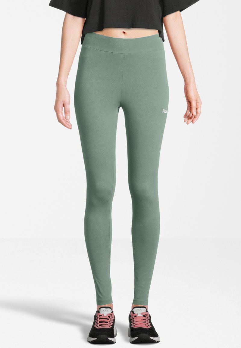 Vrouw in hoge taille saliegroene leggings en zwarte sportieve schoenen met roze veters, standing tegen een witte achtergrond.