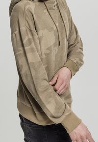 Béžová maskáčová mikina s kapucí na šňůrku, dlouhými rukávy a žebrovanými manžetami. Materiál vypadá měkce a má strukturu.