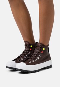 Hnědé kožené vysoké boty s bílou pryžovou podrážkou, vybavené barevnými dírkami na tkaničky a texturovanou podrážkou pro lepší grip.