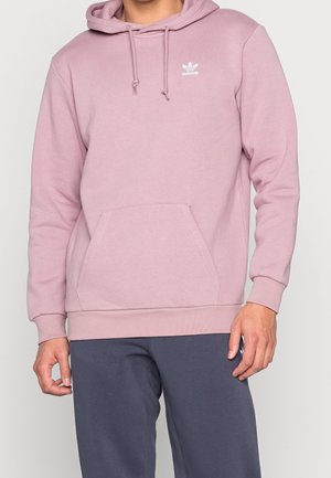 Person iført en lys lilla Adidas hættetrøje med frontlomme og snørede hætte, kombineret med mørkeblå bukser.