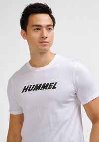 Hvid kortærmet T-shirt lavet af bomuld, med et tydeligt sort "HUMMEL" logo på tværs af brystet. Glat tekstur.