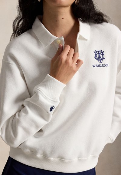 Femme portant un polo-sweatshirt blanc de Wimbledon avec col à boutons, logos brodés sur la poitrine et la manchette, tenant le col de la main.