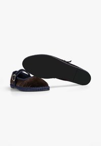 Zapato de ante marrón con suela de tejido azul, que presenta una correa con un acento metálico circular. La parte inferior es lisa y de color negro sólido.