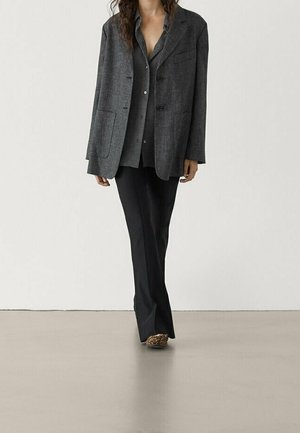 Femme portant un blazer gris foncé, une chemise grise boutonnée, un pantalon noir et des chaussures imprimé léopard, debout sur un sol lisse avec un fond uni.