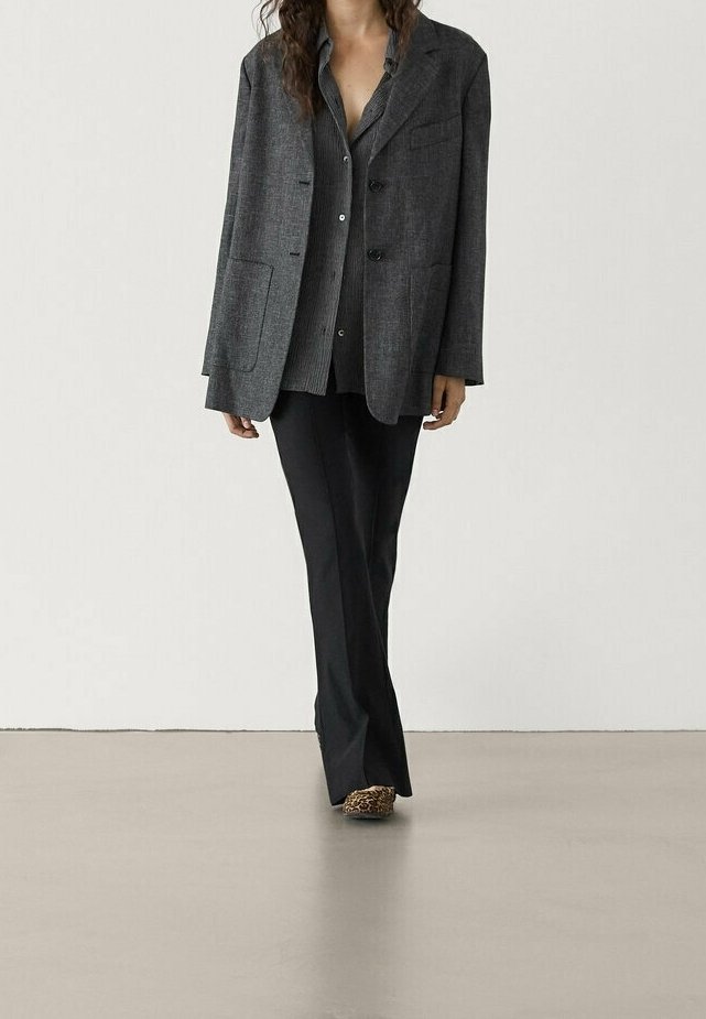 Femme portant un blazer gris foncé, une chemise grise boutonnée, un pantalon noir et des chaussures imprimé léopard, debout sur un sol lisse avec un fond uni.