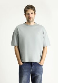 Camiseta de gran tamaño en gris claro, fabricada con una tela suave, con cuello redondo y mangas cortas, que presenta un pequeño logo bordado en el pecho.