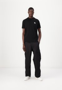 Tricou negru din bumbac cu un mic logo alb pe pieptul stâng, asortat cu pantaloni cargo negri cu buzunare laterale și adidași albi.