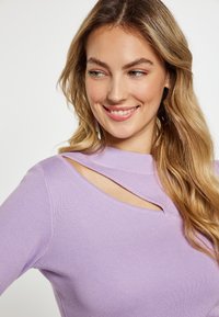 Lila Pullover mit geribbelter Struktur und einzigartigem Ausschnitt-Design am Hals. Das Modell hat welliges Haar und trägt hängende Ohrringe.