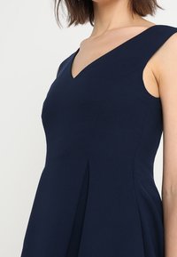 Námořnické modré šaty s výstřihem do V, bez rukávů a plisovanou sukní. Vyrobeno z hladké tkaniny; má šikmé ramínka.
