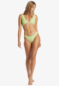 Kvinna med blont hår som bär en ljusgrön ribbad bikini, går barfota mot en vit bakgrund, tittar ner och åt sidan.