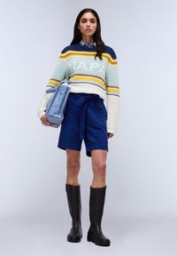 Gebreide multikleurige trui met de tekst "NAPA", gecombineerd met marineblauwe shorts en zwarte rubberen knielange laarzen. Houdt een lichtblauwe tas vast.