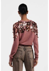 Maglione rosa con un tessuto testurizzato e dettagli di paillettes sulle spalle. Indossato con jeans a vita alta di colore marrone chiaro. Vista posteriore.