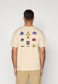 Μπεζ T-shirt με εικονογραφημένα εξωτερικά ρούχα, υποδήματα και εξοπλισμό σε διάφορα χρώματα στην πλάτη, με ετικέτα λογότυπου στο κέντρο.