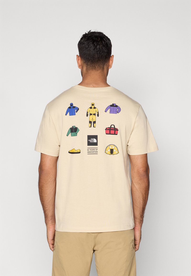 Μπεζ T-shirt με εικονογραφημένα εξωτερικά ρούχα, υποδήματα και εξοπλισμό σε διάφορα χρώματα στην πλάτη, με ετικέτα λογότυπου στο κέντρο.