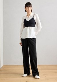 Cropped bralette nera sopra una camicia bianca con bottone e righe verticali, abbinata a pantaloni neri a gamba larga. La modella indossa scarpe slip-on bianche.