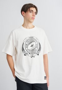 Weißes Baumwoll-T-Shirt mit einem großen schwarzen Grafikdruck, der einen Rugbyball und Textelemente zeigt, kurze Ärmel und eine lockere Passform.