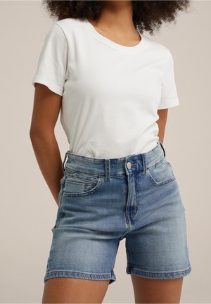 Femme portant un t-shirt blanc uni à manches courtes rentré dans un short en jean bleu clair taille haute, sur fond neutre.