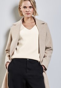 Cappotto in lana beige con rever, sopra un maglione a coste color crema, abbinato a pantaloni neri a vita alta. L'outfit è su misura e aderente.