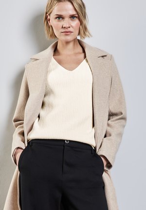 Manteau en laine beige avec des revers crantés sur un pull côtelé crème, associé à un pantalon noir taille haute. La tenue est ajustée et bien coupée.
