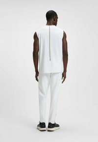 Witte mouwloze top met een verticale streepdetail op de rug, gecombineerd met witte broek en zwarte sneakers. Gladde textuur, modern ontwerp.