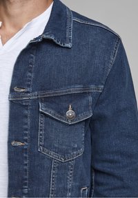Denim jacket in dark blue with a classic collar, chest pocket, copper button details, and subtle distressed accents. White t-shirt underneath.