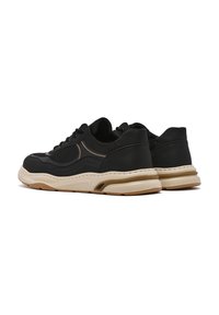 Un par de zapatillas negras de corte bajo con suelas de goma beige y marrón, mostradas desde la parte trasera y lateral sobre un fondo blanco.