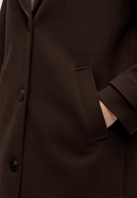Gros plan d'un manteau en laine brun foncé avec poche à rabat cousue et boutons noirs, montrant une main partiellement dans la poche.