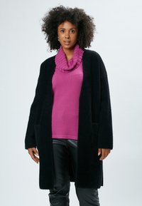 Cardigan nero e morbido con apertura frontale e tasche laterali, indossato su un maglione rosa a collo alto con texture e pantaloni neri aderenti.