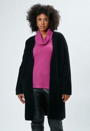 Schwarze, flauschige Strickjacke mit offenem Vorderteil und seitlichen Taschen, getragen über einem rosa, strukturierten Kragenpullover und schwarzen, eng anliegenden Hosen.