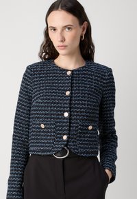 Veste en tweed bleu marine et bleu avec un motif texturé, dotée de cinq boutons dorés, poches horizontales et d'un col rond.
