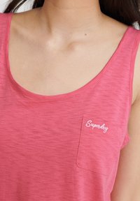 Lyserød ærmeløs tanktop i blødt stof med en rund halsudskæring og en lille lomme med "Superdry" logo i hvid brodering.