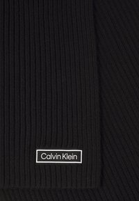 Zwart ribgebreid stof met een klein rechthoekig Calvin Klein logo label in wit en zwart nabij de onderrand.