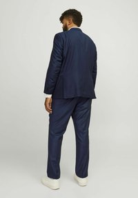 Navy blue suit with structured shoulders, smooth fabric, and slim fit. White shirt cuffs visible. White sneakers complete the look.