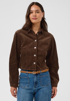 Femme portant une chemise en velours côtelé marron avec des boutons argentés, un jean taille haute bleu et une ceinture beige, debout devant un fond uni.