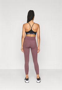 Μαύρο αθλητικό σουτιέν με ρυθμιζόμενους ιμάντες, που διαθέτει λευκή λωρίδα με λογότυπο. Μωβ κολάν ψηλής μέσης με ομαλή υφή, συνδυασμένα με μαύρα αθλητικά παπούτσια.