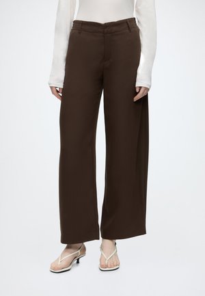 Personne portant un pantalon marron taille haute à jambes larges, associé à un haut blanc à manches longues et des sandales blanches à brides fines et petits talons kitten heel.