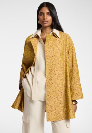 Mujer con un abrigo amarillo de textura floral sobre una blusa crema y pantalones cargo, con pendientes de aro dorados y pulseras, de pie con las manos en los bolsillos.