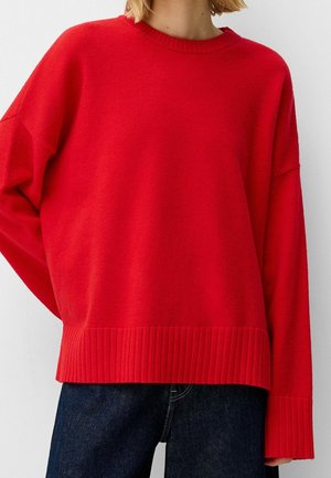Frau trägt einen locker sitzenden roten Langarmpullover mit gerippten Bündchen und Saum, kombiniert mit dunkelblauen Jeans.