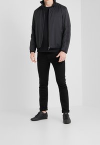 Muž v černé zipové bundě, černém tričku, černých úzkých džínách a černých teniskách na světlém pozadí.