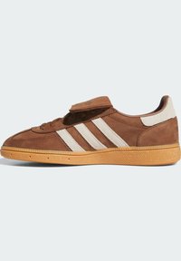 Hnedé semišové tenisky s bielymi prúžkami, gumovou podrážkou a detailom prehnutého jazyka, zobrazené z vonkajšej strany na svetlom pozadí.