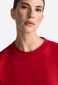 Červená mikina s okrúhlym výstrihom, s textúrovanou tkaninou a rebrovanými manžetami. Detailný pohľad na výstrih a tvár.