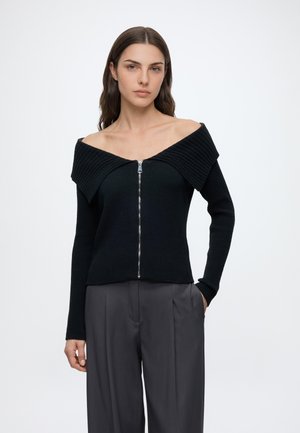 Kvinde med langt brunt hår iført en sort off-shoulder lynlås trøje og mørkegrå vidde bukser, står mod en hvid baggrund.