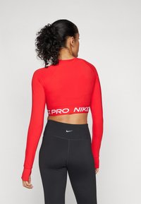 Crvena dugosleeve skraćena majica s bijelim natpisima "PRO" i "NIKE" na pojasu, uparen s crnim tajicama visokog struka. Glatka tekstura.