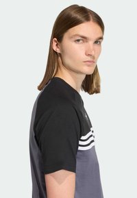 Tričko s krátkymi rukávmi v čiernej a sivej farbe s bielymi vodorovnými pruhmi a okrúhlym výstrihom. Hladká bavlnená textúra.