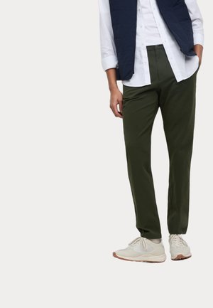 Mężczyzna ubrany w oliwkowozielone spodnie, białe trampki, białą koszulę i granatową kamizelkę bez rękawów, stoi z jedną ręką w kieszeni.