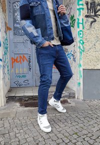 Džínová bunda s patchworkem v různých odstínech modré, béžové tričko, úzké tmavé džíny a bílé kotníkové tenisky se šedými akcenty.