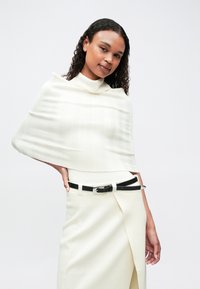 Giovane donna con capelli ricci che indossa un top in maglia a coste bianco e una gonna, accessoriate con una cintura nera, in posa di fronte a uno sfondo neutro.